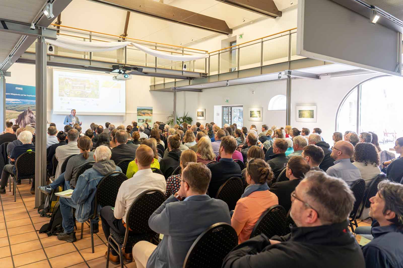 Ein überfüllter Konferenzsaal mit Teilnehmern, die einem Referenten und einem großen Bildschirm folgen.