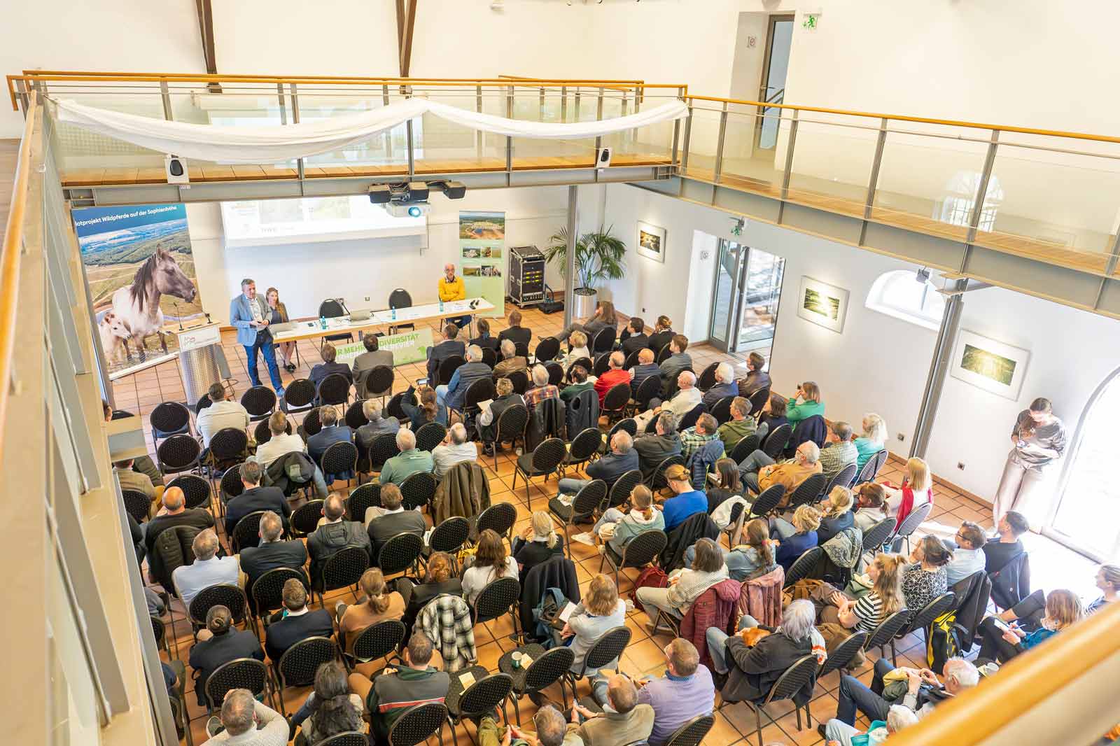 Ein Publikum sitzt in einem Konferenzsaal und schaut aufmerksam einer Präsentation vorne zu, mit modernem Dekor.
