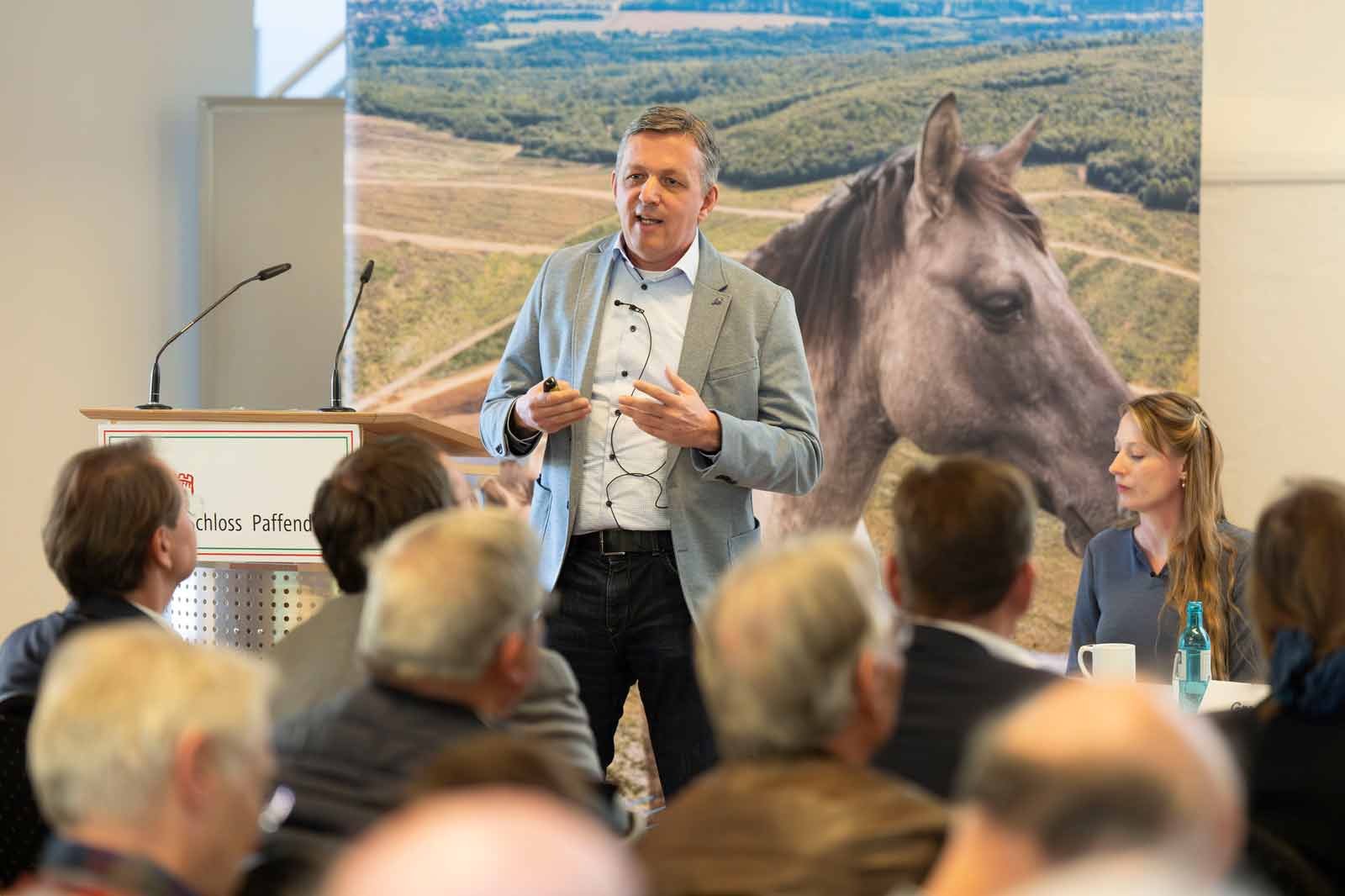 Ein Redner präsentiert auf einer Konferenz vor einem Hintergrund mit einem Pferd. Die Teilnehmer sind in die Sitzung vertieft.
