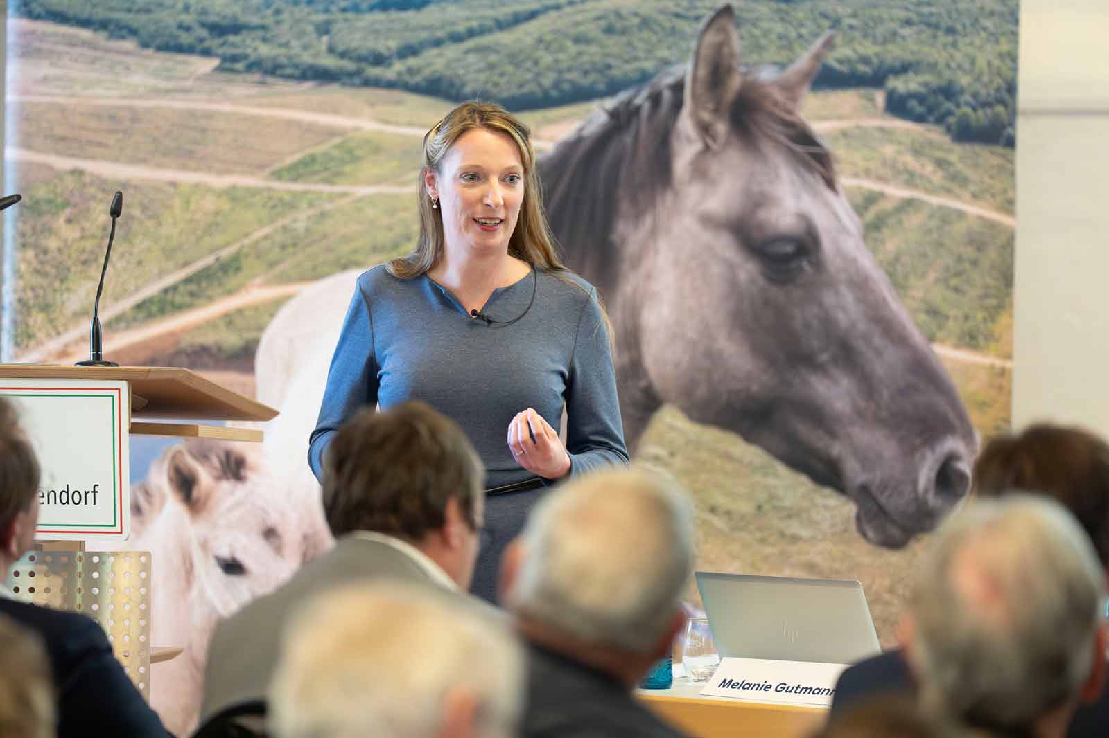Eine Frau spricht an einem Podium mit einem Pferdebild hinter sich, während das Publikum aufmerksam zuhört.