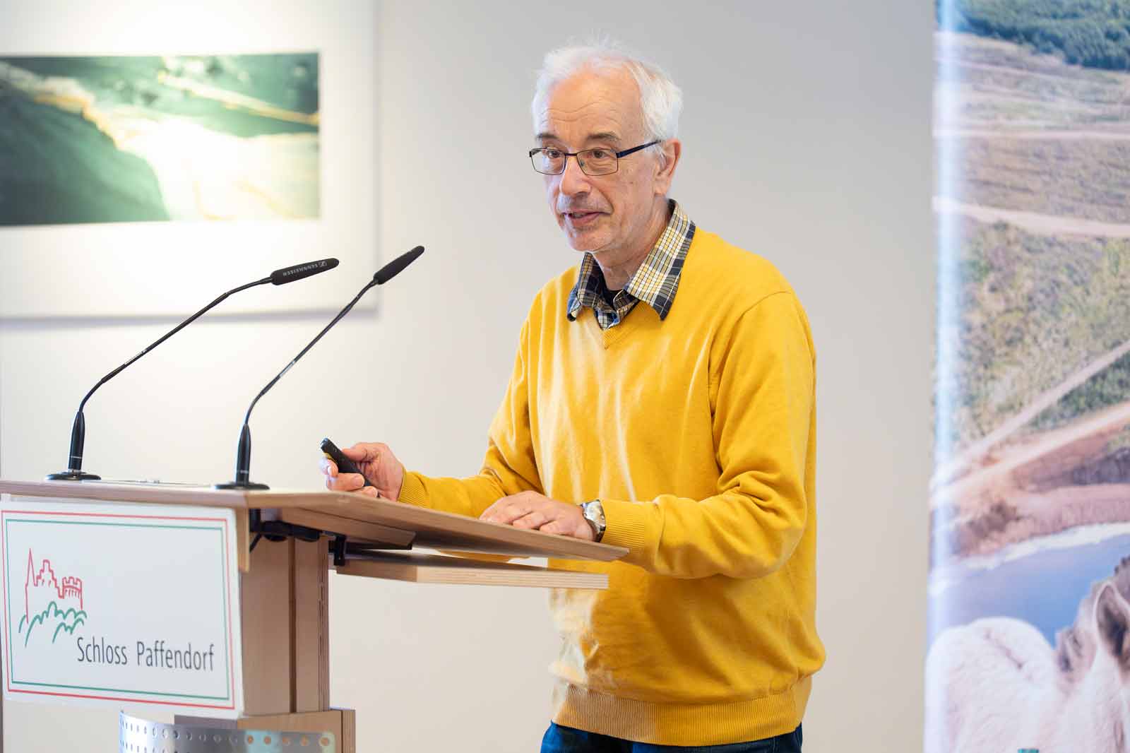 Ein Redner in einem gelben Pullover präsentiert an einem Podium mit Mikrofonen vor einer Wand mit einem Naturbanner.