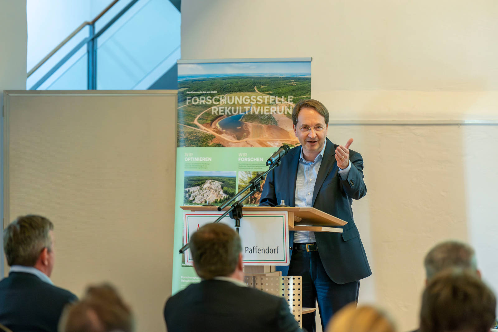 Ein Redner gestikuliert an einem Podium während einer Präsentation, im Hintergrund sind Slogans und Bilder zur Umweltforschung zu sehen.