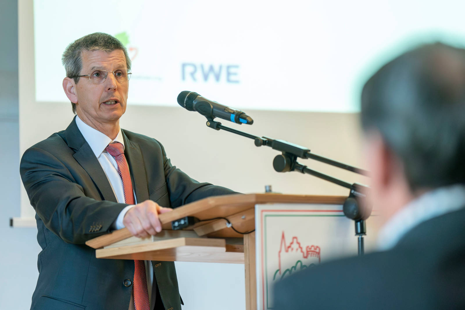 Ein Redner an einem Podium, der präsentiert, mit einem Mikrofon davor. Im Hintergrund sind Unternehmenslogos zu sehen.