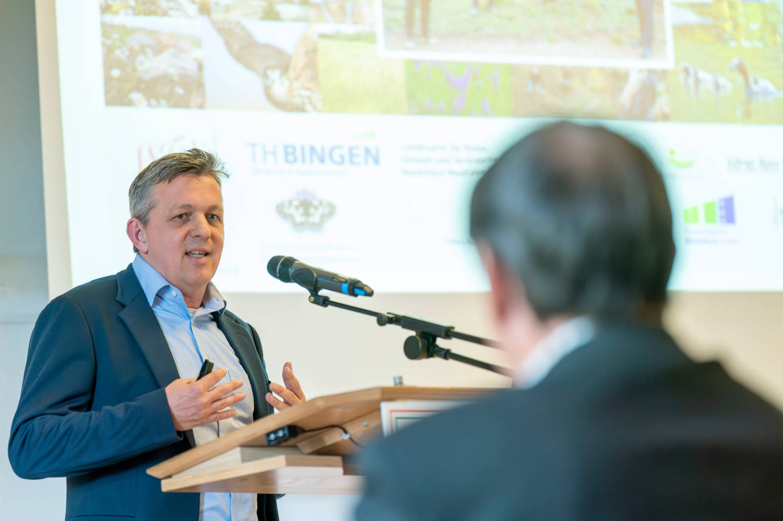 Ein Redner präsentiert auf einer Konferenz und gestikuliert in Richtung eines Bildschirms mit verschiedenen Bildern und Logos in einem professionellen Umfeld.