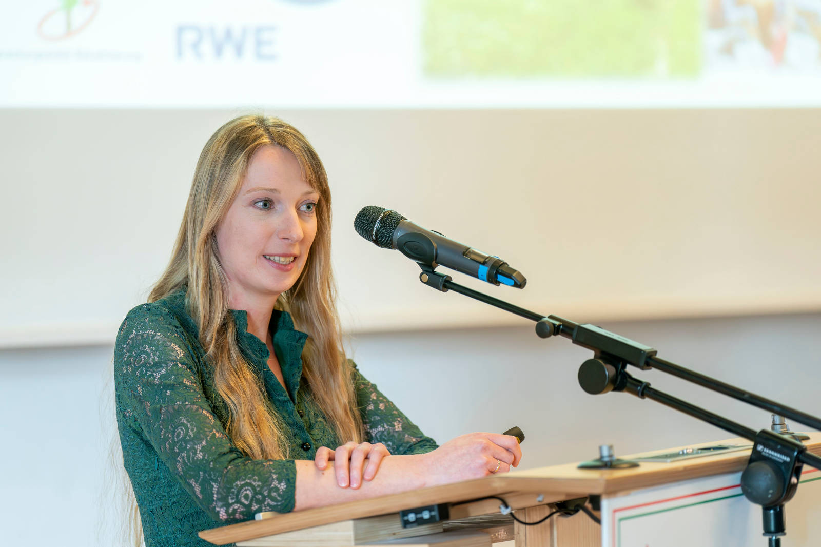 Eine Frau steht an einem Rednerpult mit einem Mikrofon und hält eine Präsentation vor verschwommenem Hintergrund mit verschiedenen Logos.