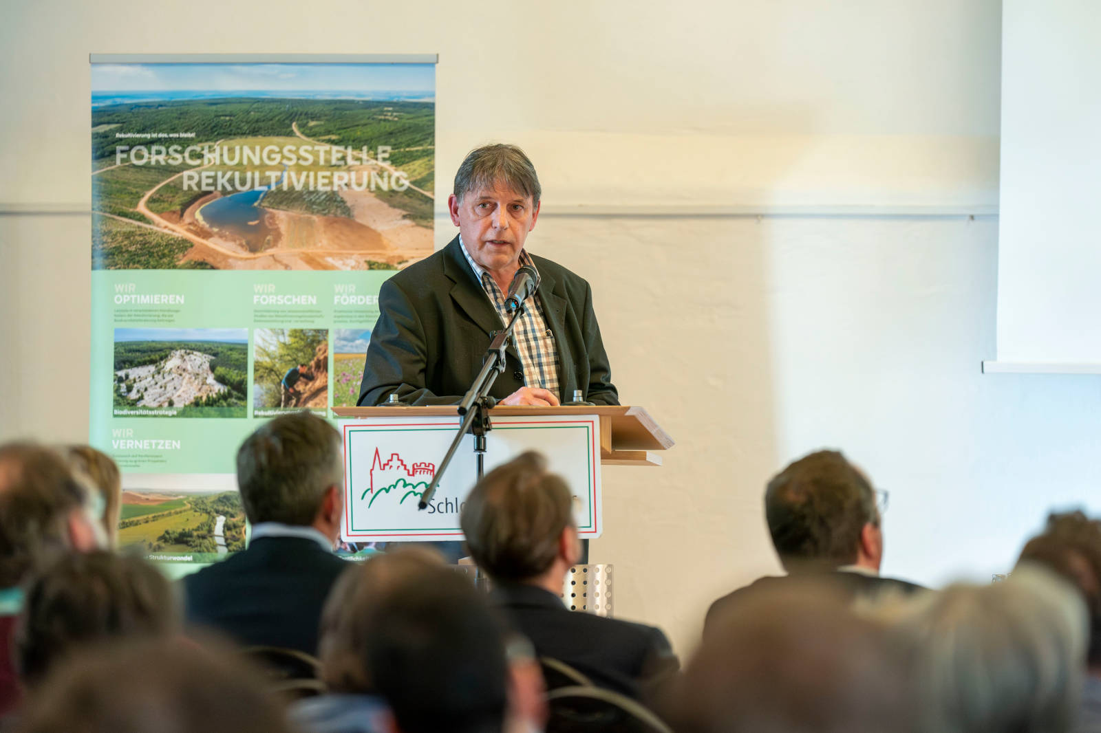 Ein Redner präsentiert auf einer Konferenz vor einem Banner über Forschung und Rekultivierung.