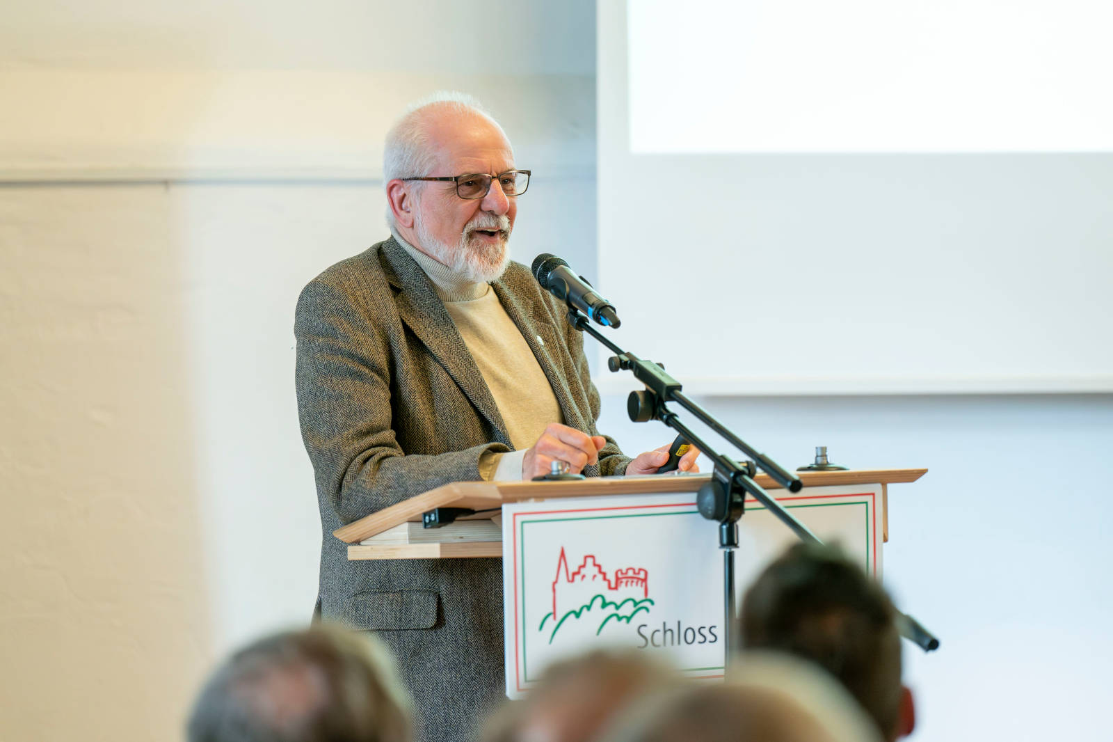 Ein Redner steht an einem Podium und präsentiert auf einer Konferenz. Im Hintergrund wird eine Präsentationsfolie angezeigt.