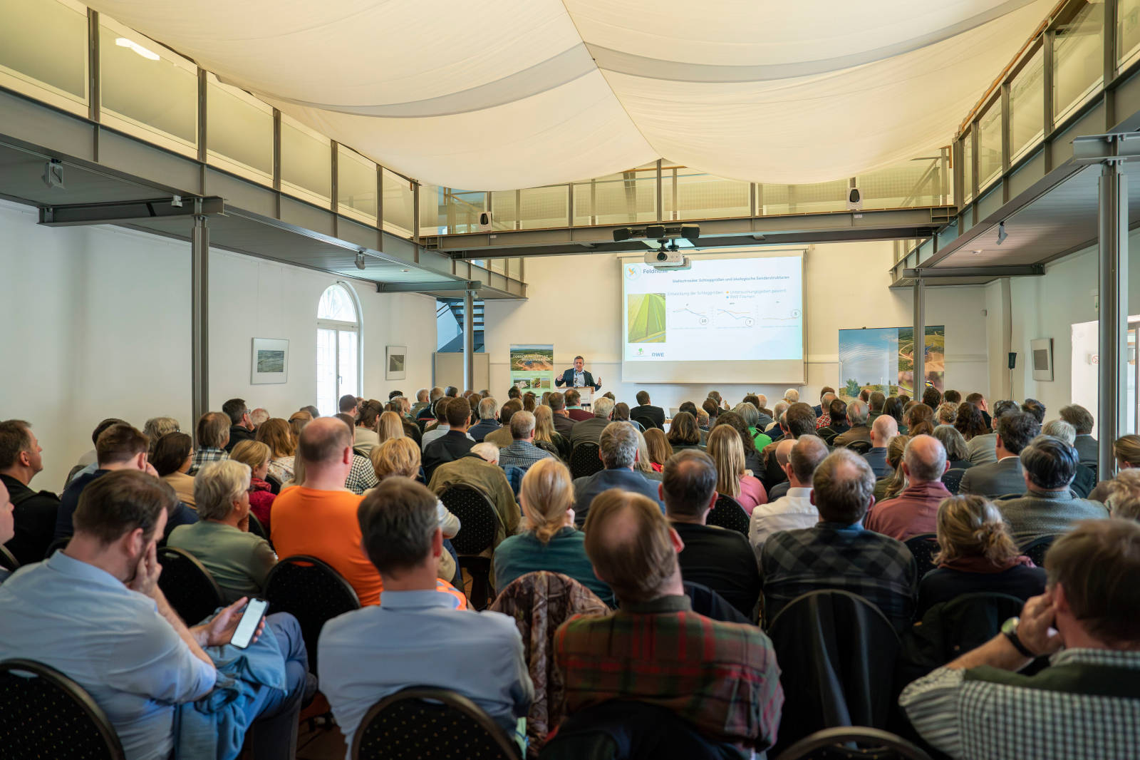Ein voller Seminarraum mit Teilnehmern, die einem Redner zuhören, der Informationen auf einem großen Bildschirm präsentiert.