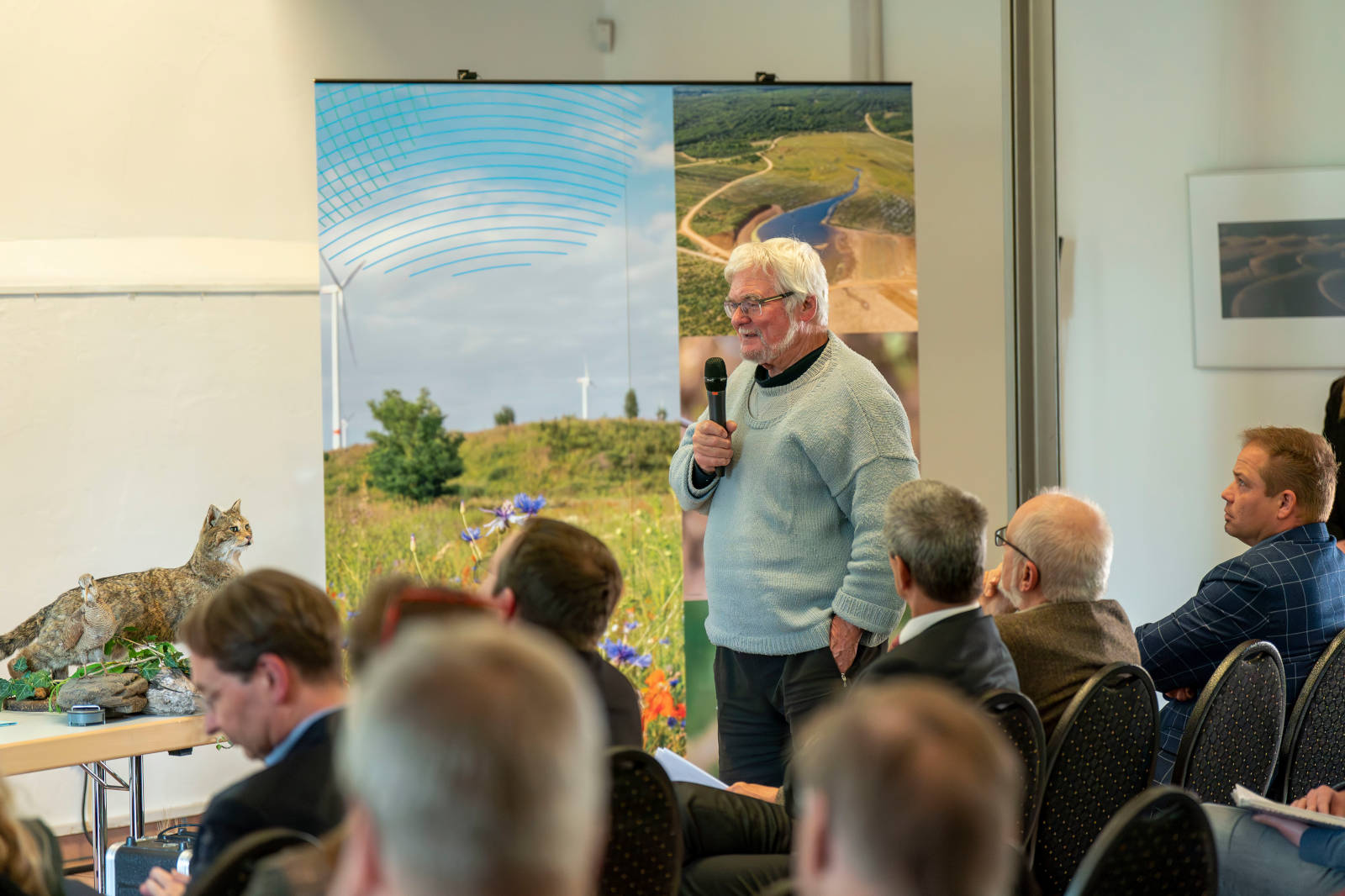 Ein Sprecher präsentiert bei einer Veranstaltung, mit einem Luchs auf einem Tisch und einem Naturhintergrund.