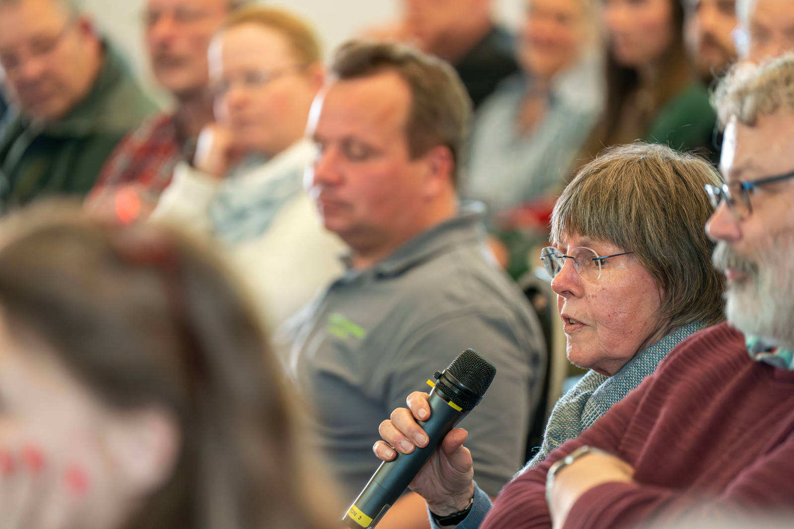Eine Menschenmenge bei einer Veranstaltung. Eine Person hält ein Mikrofon und zeigt damit, dass sie sprechen möchte.