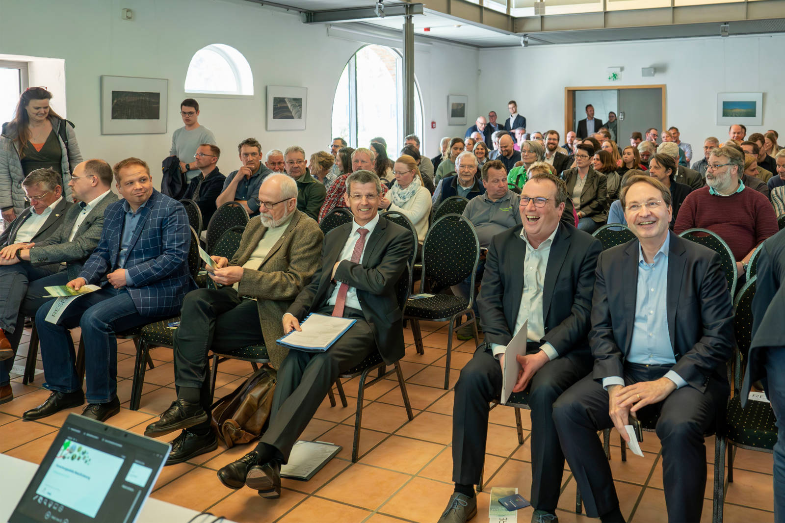 Eine große Zuhörerschaft sitzt in einem Besprechungsraum und hört aufmerksam einer Präsentation zu. Einige Personen halten Unterlagen.
