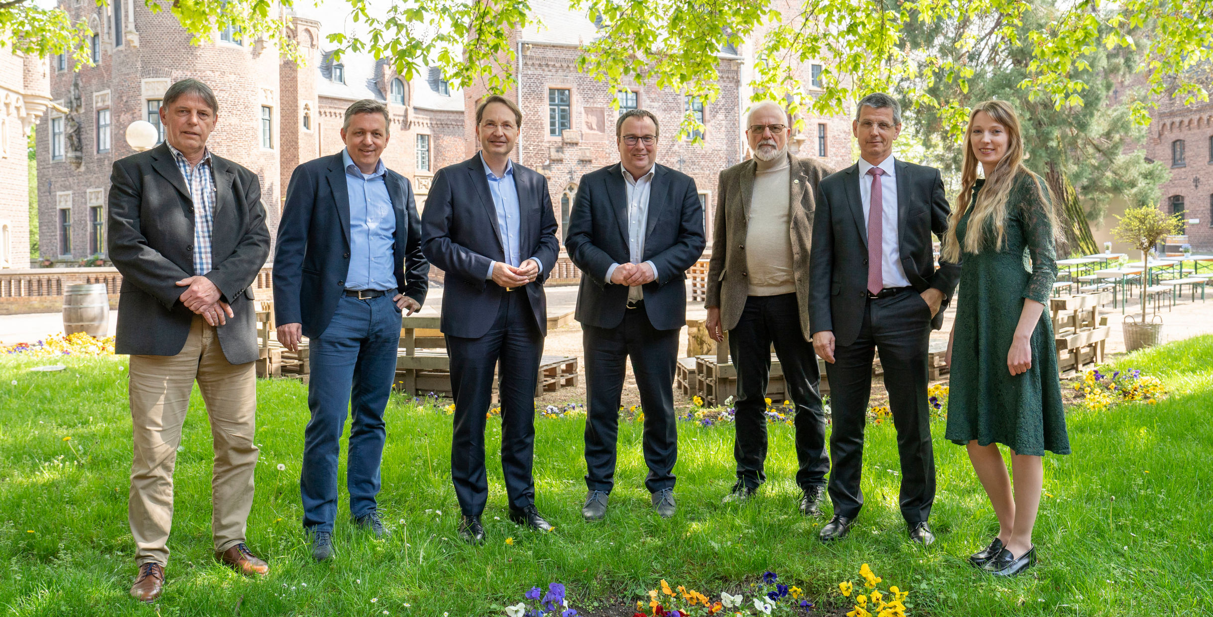 Eine Gruppe von sieben Personen steht in einem Garten mit einem historischen Gebäude im Hintergrund, umgeben von grünem Gras und Blumen.
