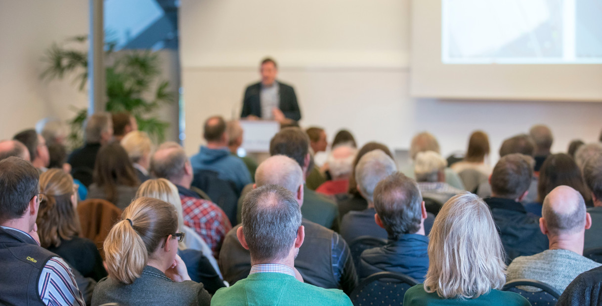 Ein diverses Publikum hört aufmerksam einem Redner während einer Präsentation in einem modernen Konferenzraum zu.